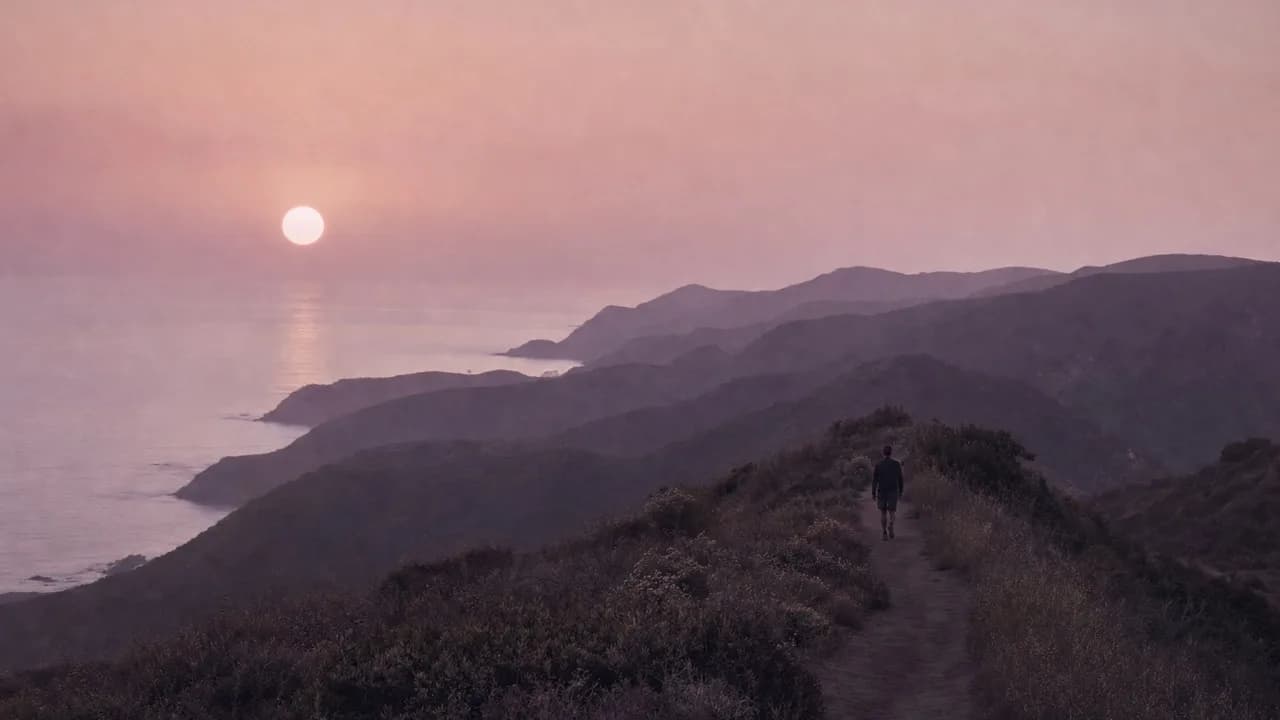 A person walking a narrow ridge trail through coastal sage at dusk, back to camera, layered hills fading into mauve haze toward the sea and a pale sun.