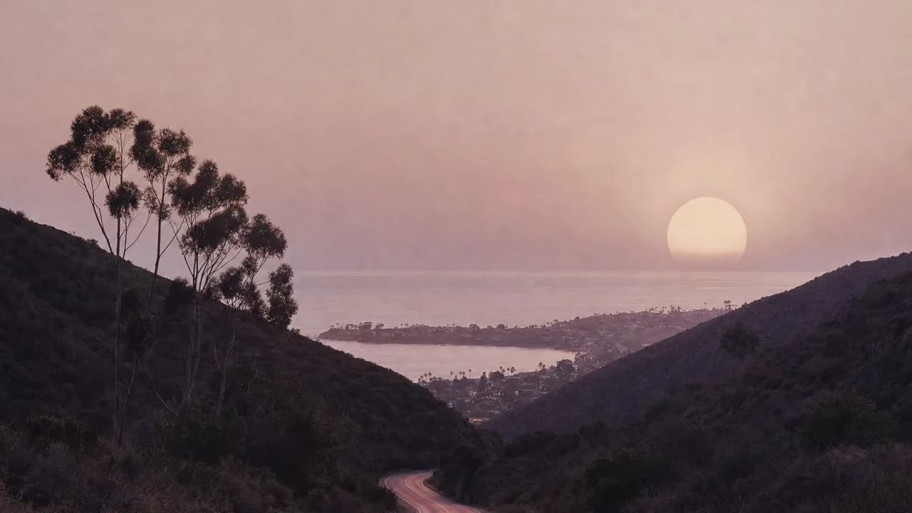 A road winding down through dark canyon hills and eucalyptus toward a coastal town at dusk, a huge pale sun over the sea.