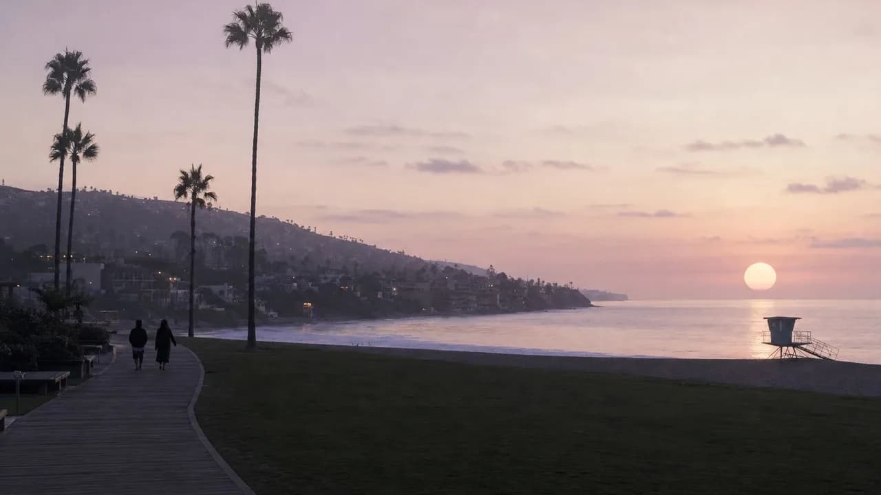 Two people walking a boardwalk past tall palms at a beachfront park in Laguna Beach at dusk, backs to camera, a lifeguard tower and a pale sun on the water.