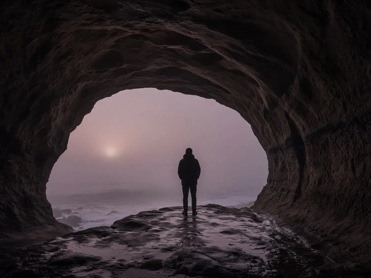A solitary figure standing at the mouth of a dark sandstone sea cave on a Laguna headland at dusk, back to camera, a pale sun beyond the opening.